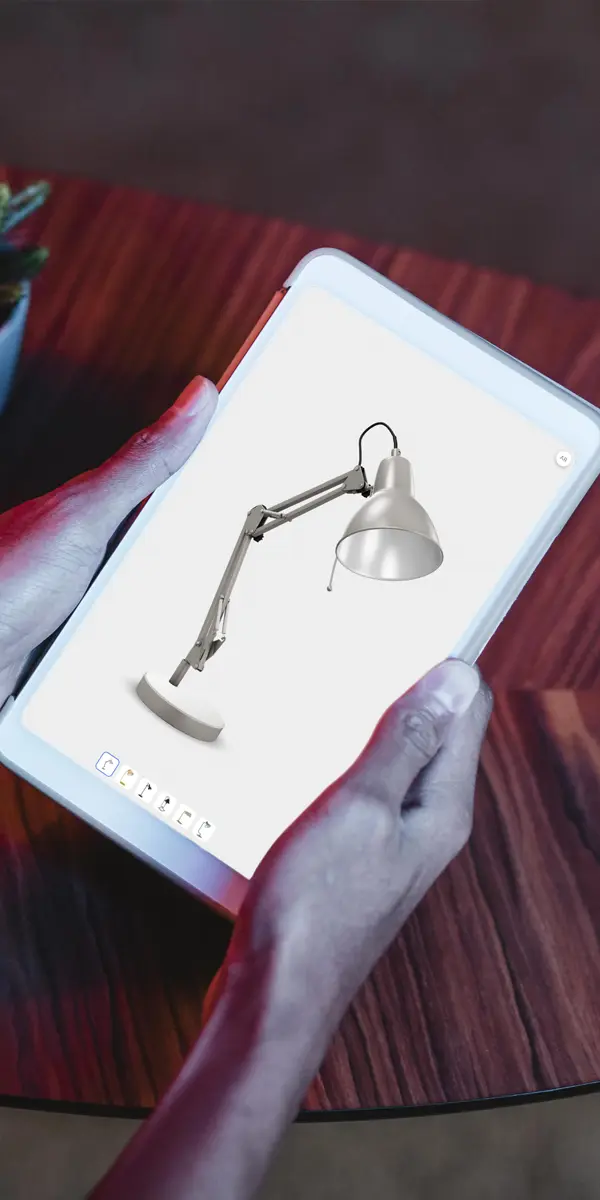 A person holding a tablet displaying an augmented reality (AR) view of a desk lamp, as if the lamp is standing on the table in 3D design